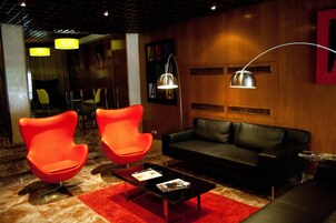 Un salón moderno con un sofá de cuero negro, dos sillas color naranja brillante, un tapete rojo y una mesa de centro con revistas.