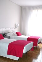Una habitación de hotel con dos camas, ropa de cama blanca, almohadas rojas, una mesita de noche, una lámpara y una ventana con cortinas traslúcidas.