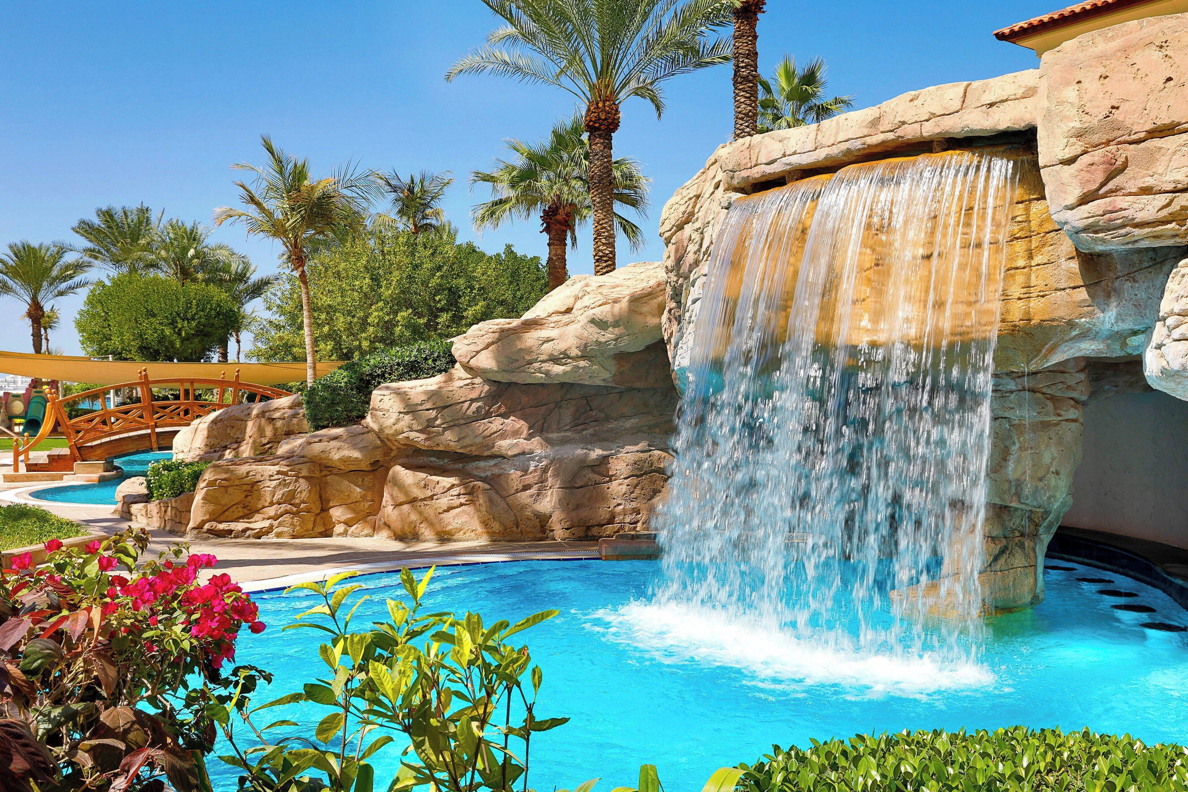 A pool with a waterfall feature, surrounded by palm trees and a bridge.