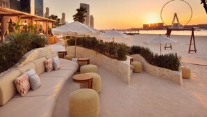 A waterfront outdoor seating area with a sofa, round tables, and umbrellas, overlooking a city skyline and a Ferris wheel.