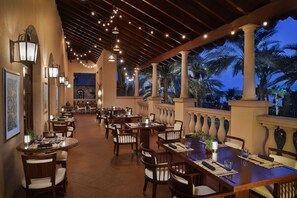 A restaurant with multiple tables and chairs, string lights, and a view of palm trees.