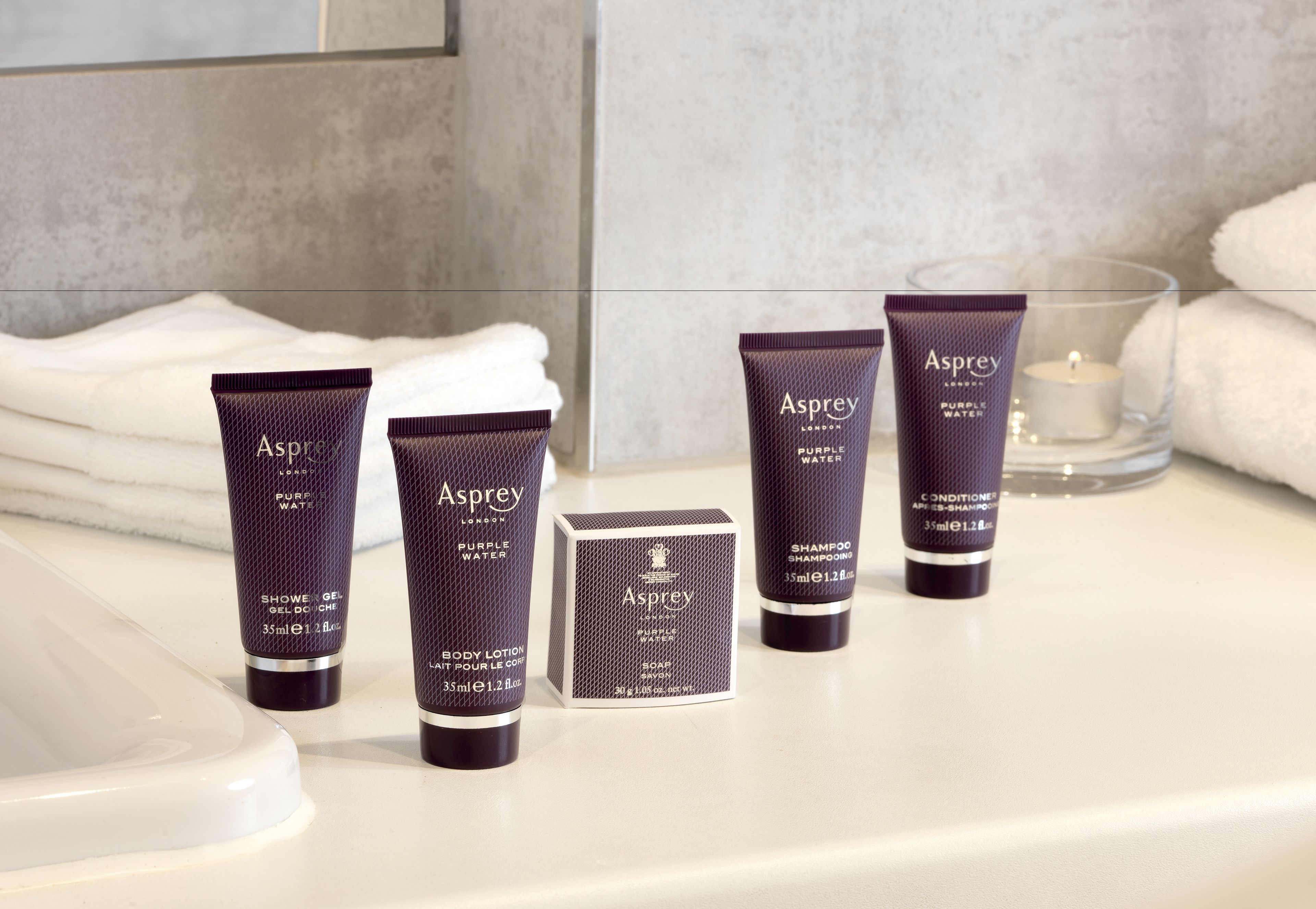 A bathroom counter with various toiletries including a purple tube of Asprey body lotion, a purple tube of Asprey shampoo, a purple tube of Asprey conditioner, and a purple tube of Asprey shower gel.