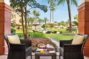 A patio with wicker chairs, a table with breakfast items, and a view of a well-maintained garden with palm trees and umbrellas.