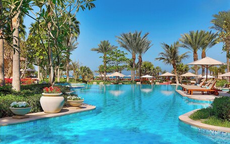 A swimming pool with lounge chairs and umbrellas, surrounded by palm trees and potted plants.