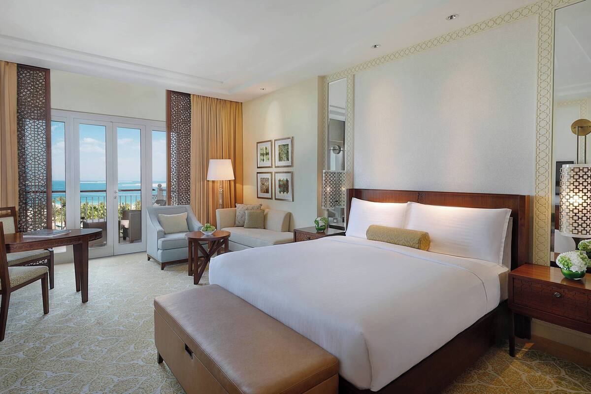 A hotel room with a large bed, a desk, a chair, and a sofa. There is a balcony with a view of the ocean.