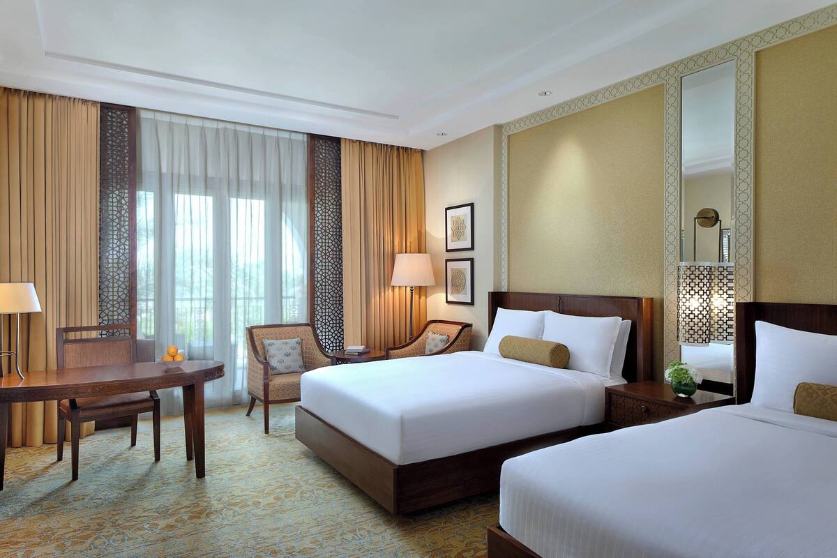 A hotel room with two beds, a desk, a chair, and a large window with curtains.