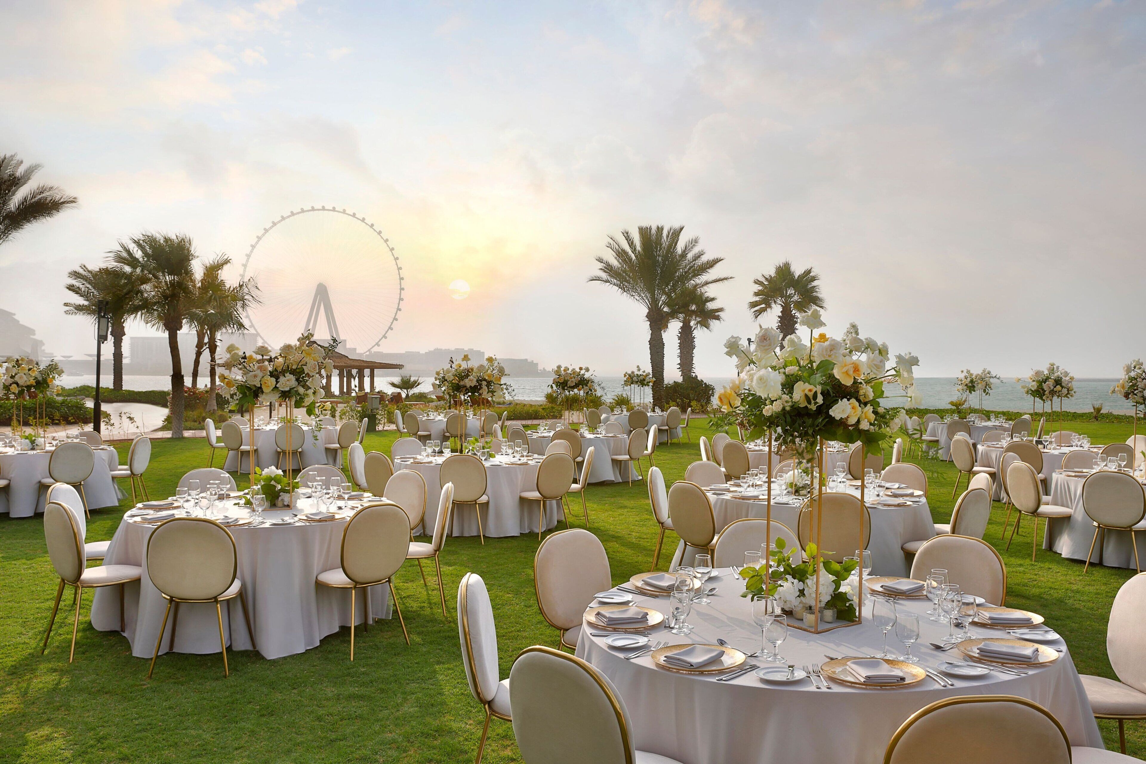 An outdoor wedding venue with round tables set up on a grassy lawn, overlooking a body of water and a Ferris wheel in the background.