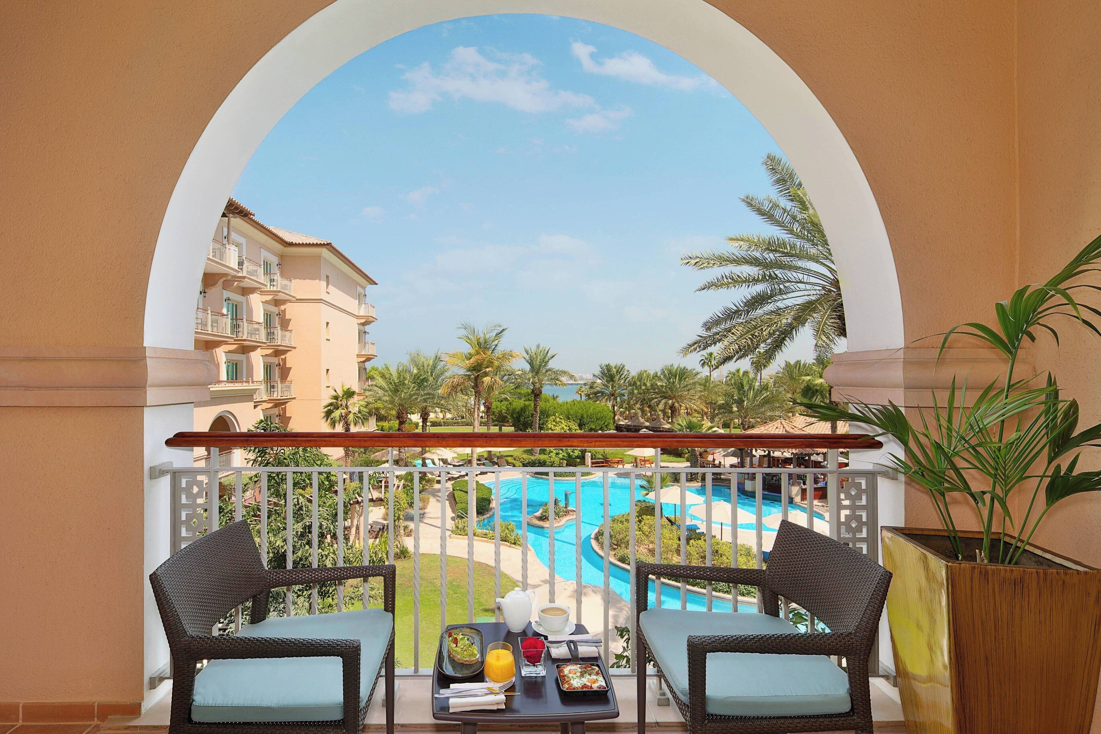 A balcony with wicker chairs, a table with breakfast items, and a view of a swimming pool and palm trees.