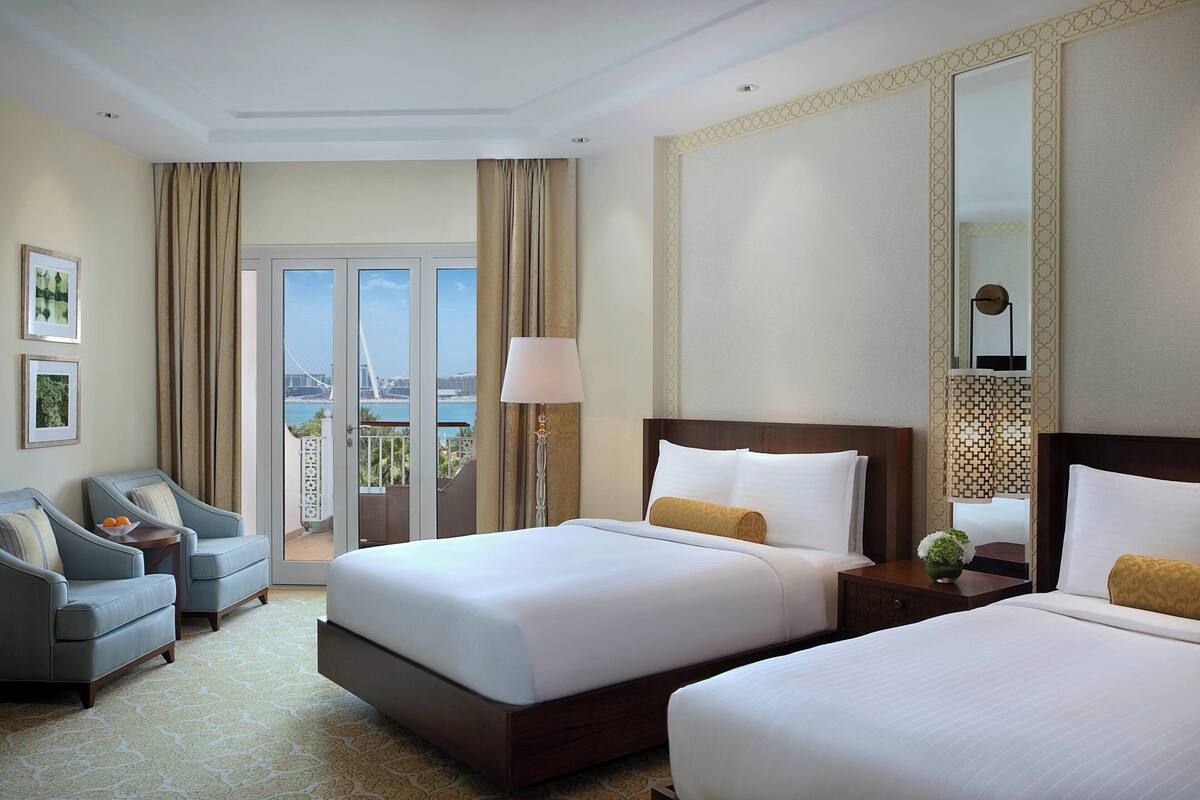 A hotel room with two beds, a sitting area with a sofa, a lamp, and a view of a bridge and cityscape.