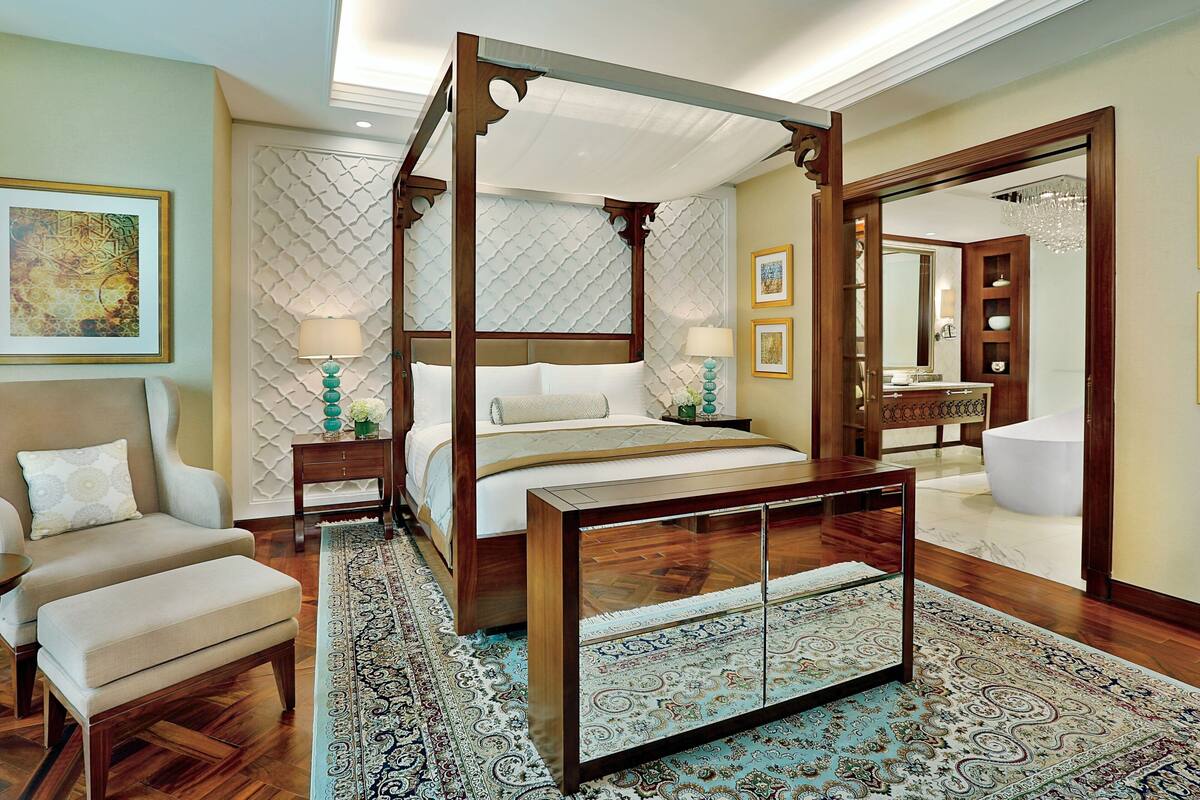 A four-poster canopy bed with a wooden frame and white bedding, a sitting area with a cushioned bench and armchair, a large mirror, and a bathroom with a bathtub and sink.
