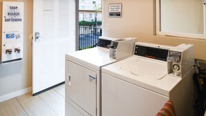 A laundry area with a front-loading washer and dryer, a sign indicating laundry services, and a view of the street outside. - Anaheim Islander Inn and Suites (Anaheim)