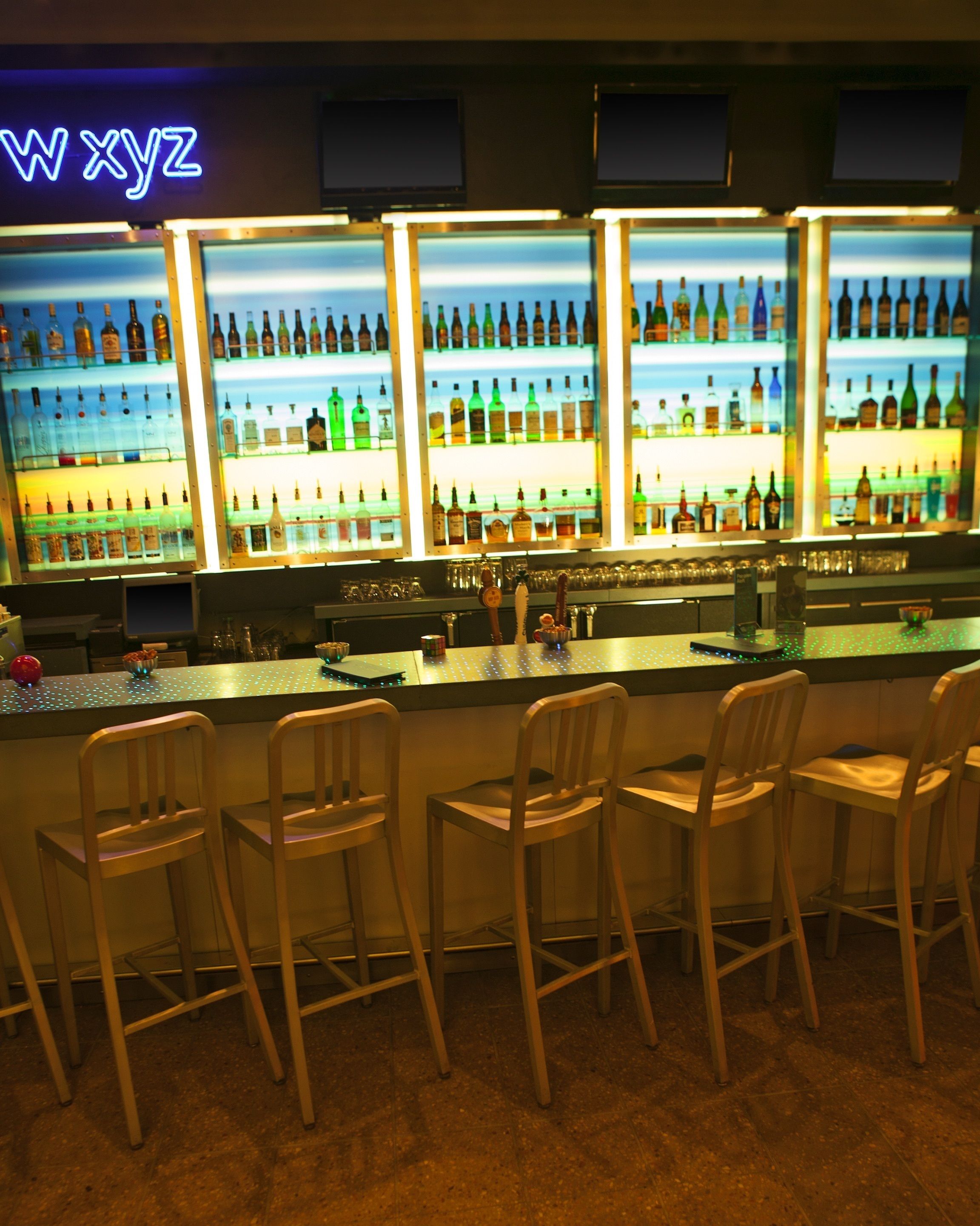 A bar area with multiple shelves of bottles, a neon sign, and bar stools.