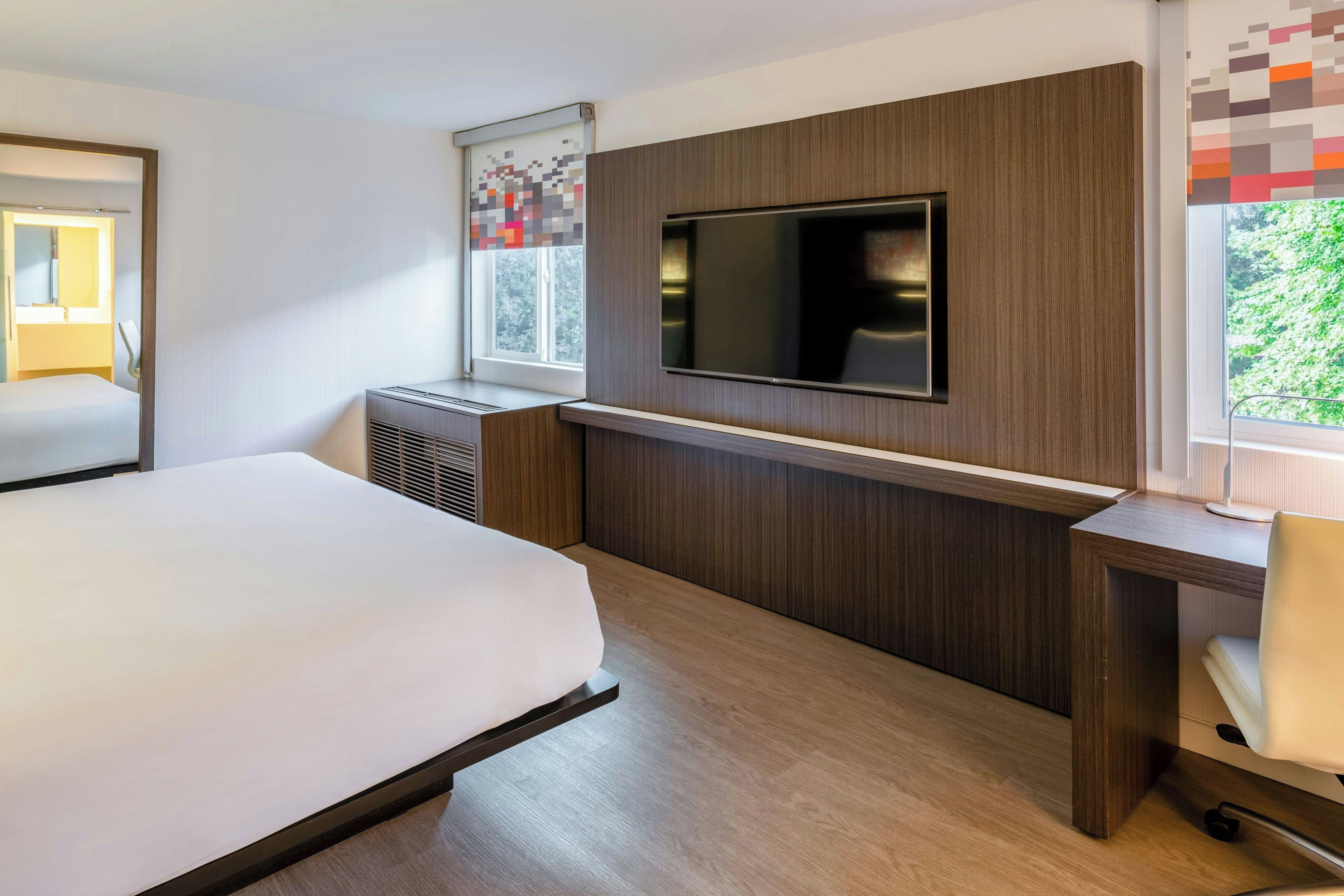 A hotel room with a large bed, a desk, a flat-screen TV mounted on the wall, and a window with a view of greenery.