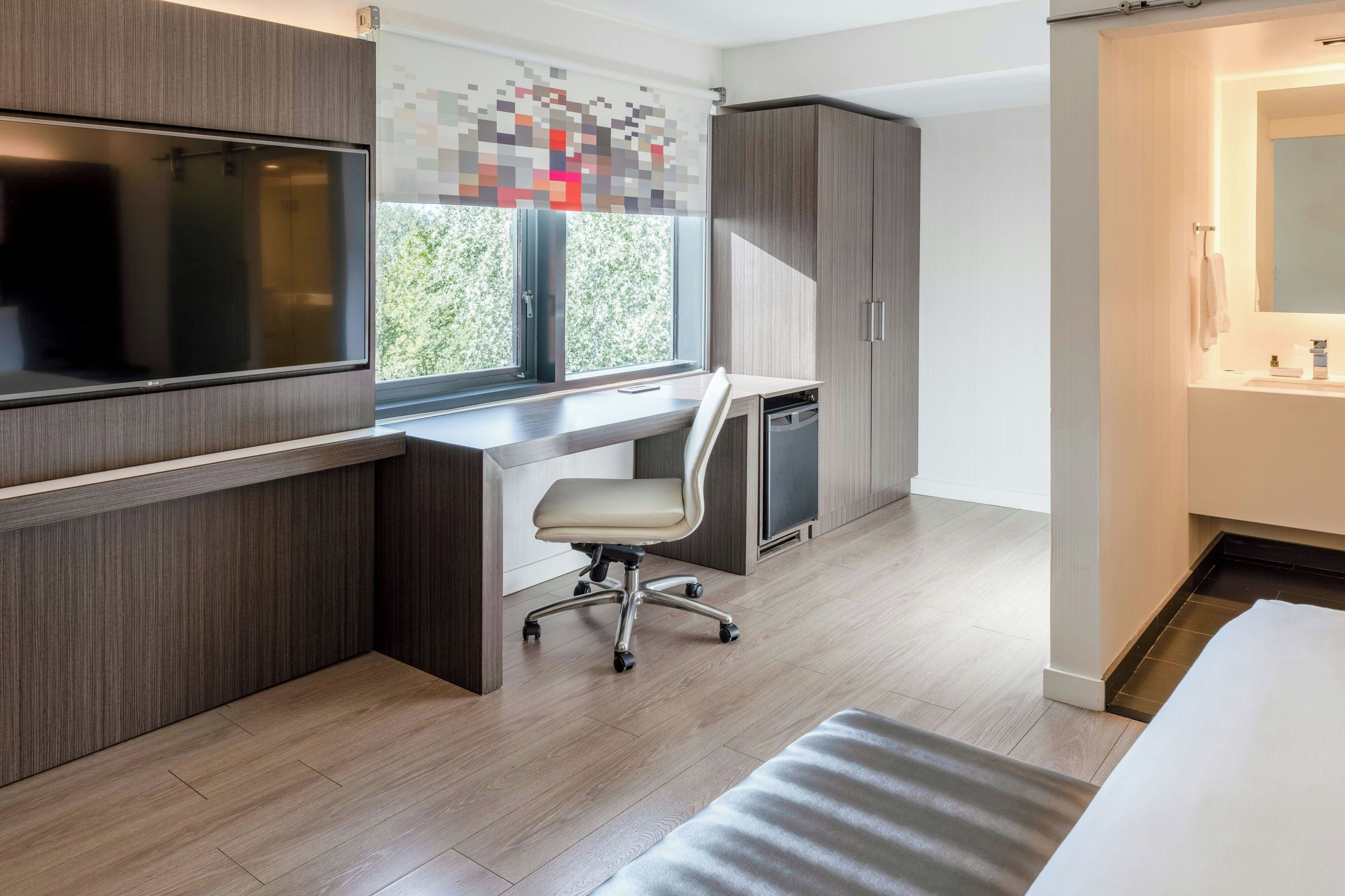 A modern bedroom with a bed, a desk with a chair, a TV, and a bathroom with a sink and mirror.