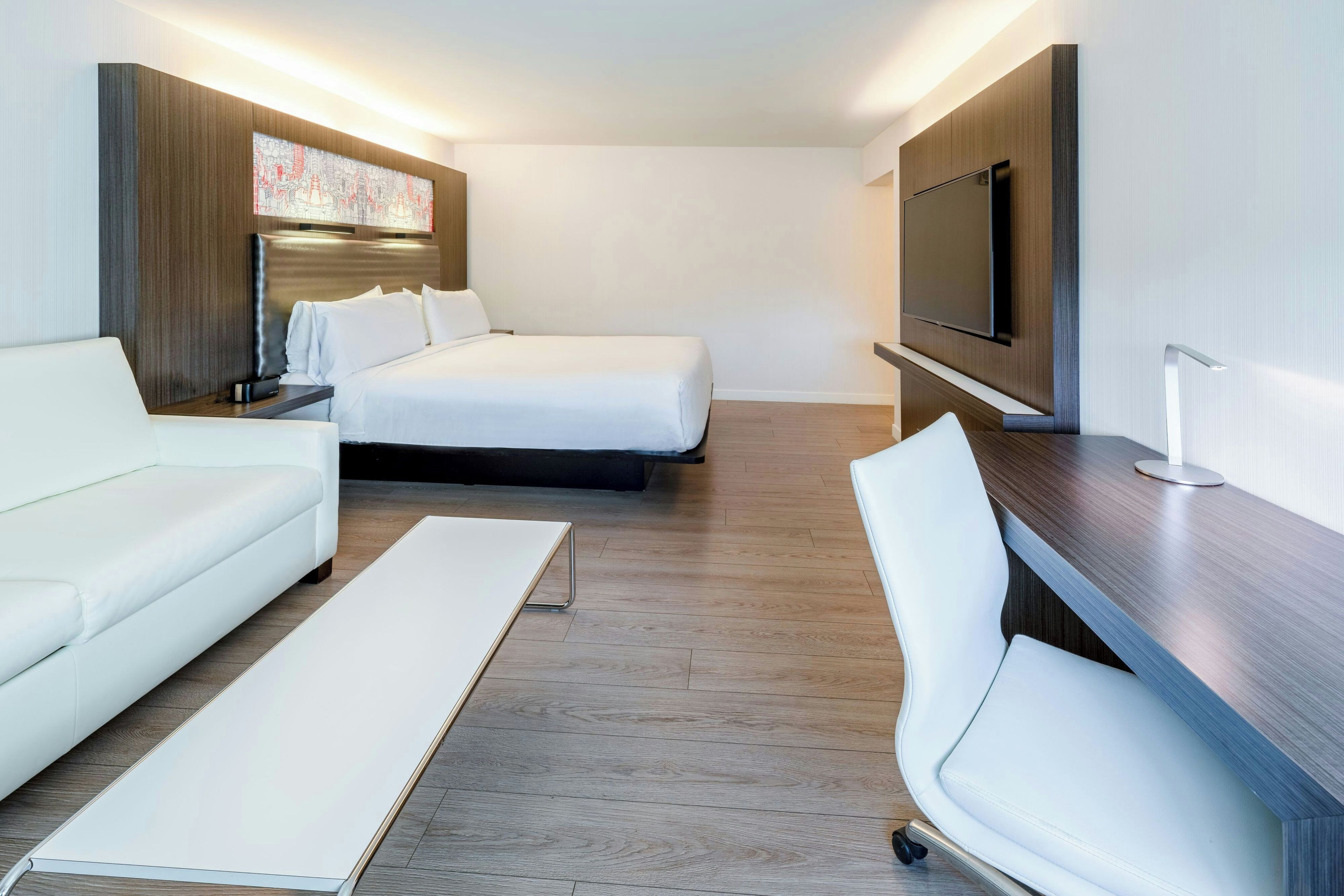 A modern hotel room with a bed, a sofa, a desk, and a flat-screen TV.