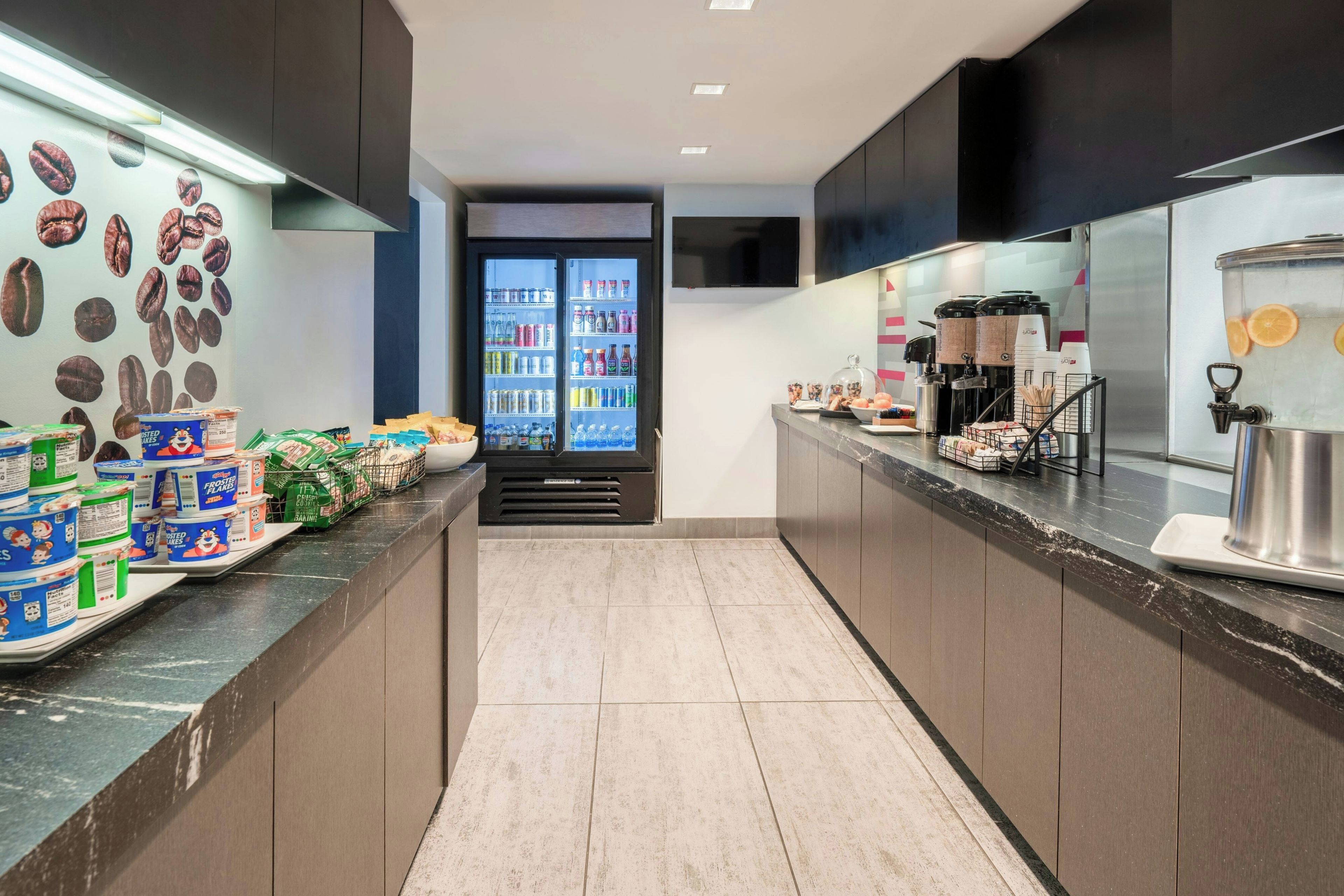 Un área de cocina moderna con una cafetera, un refrigerador lleno de bebidas y un mostrador con alimentos envasados.