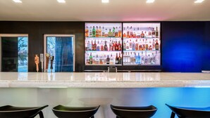 Eine Bar mit einer langen Theke und vier Hockern. Hinter der Theke befinden sich Regale mit verschiedenen Flaschen.