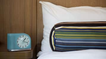 A bed with white bedding and a striped pillow, a blue clock on a wooden nightstand, and a wooden headboard.