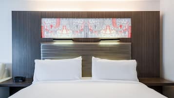 A modern hotel room with a large bed, two white pillows, a nightstand with a digital clock, and a wall-mounted artwork.