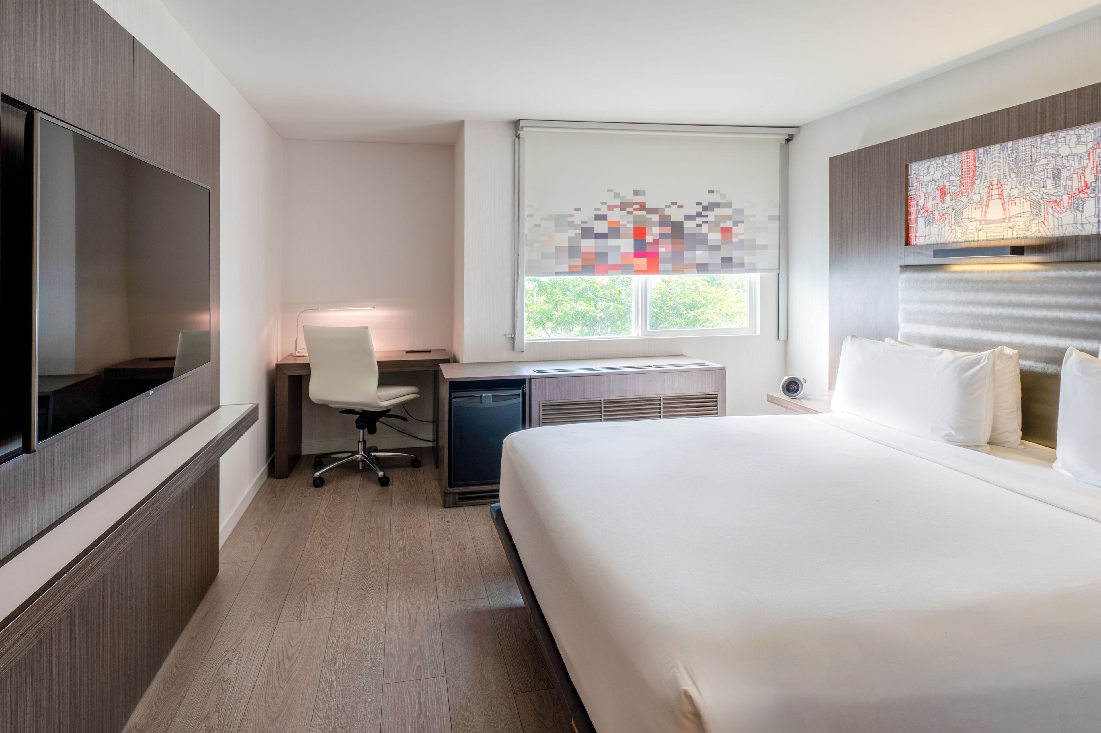 A modern hotel room with a large bed, a desk with a chair, a television, and a window with a cityscape view.