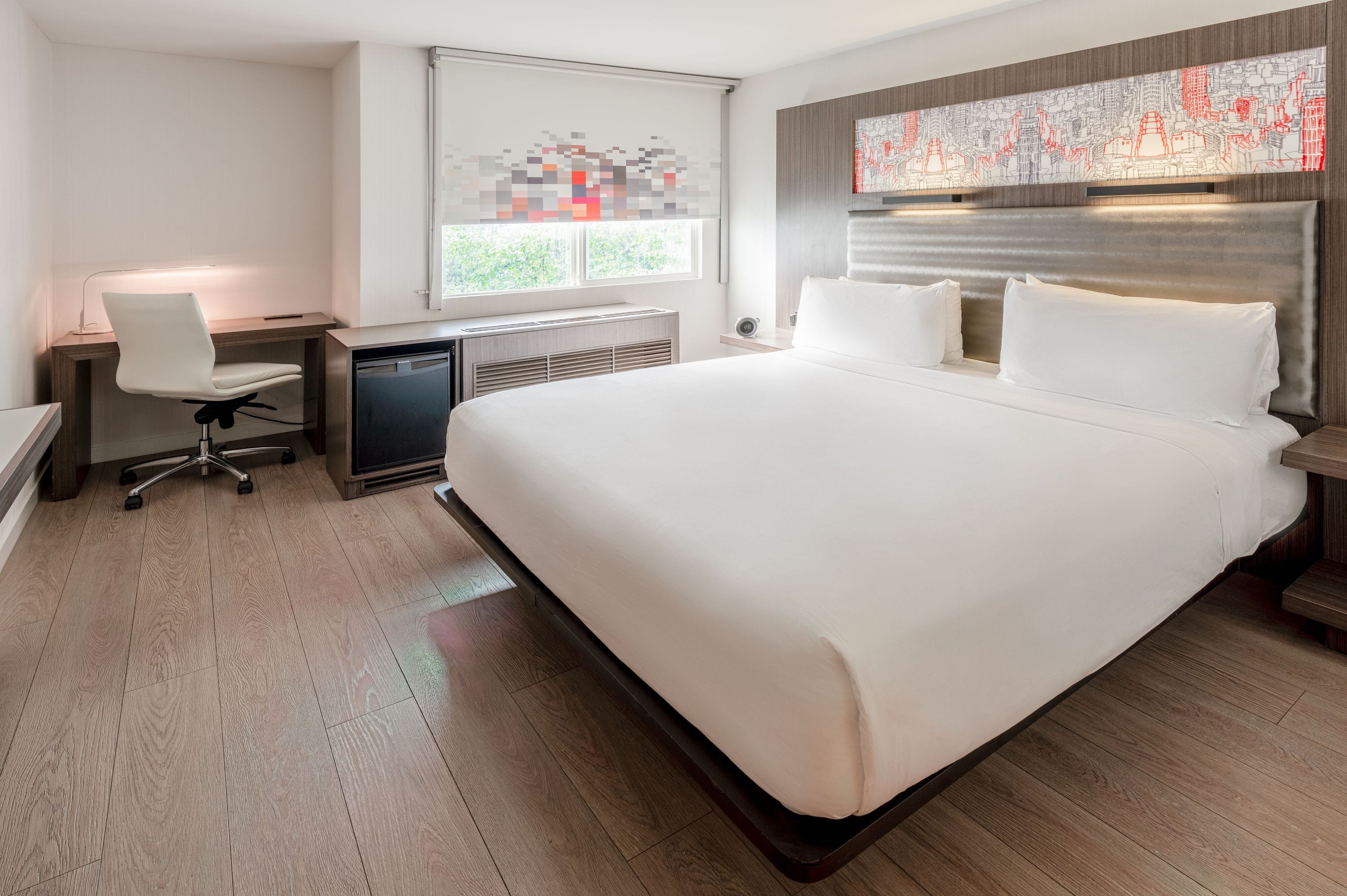 A modern hotel room with a large bed, a desk with a chair, a window with blinds, and a cityscape artwork on the wall.