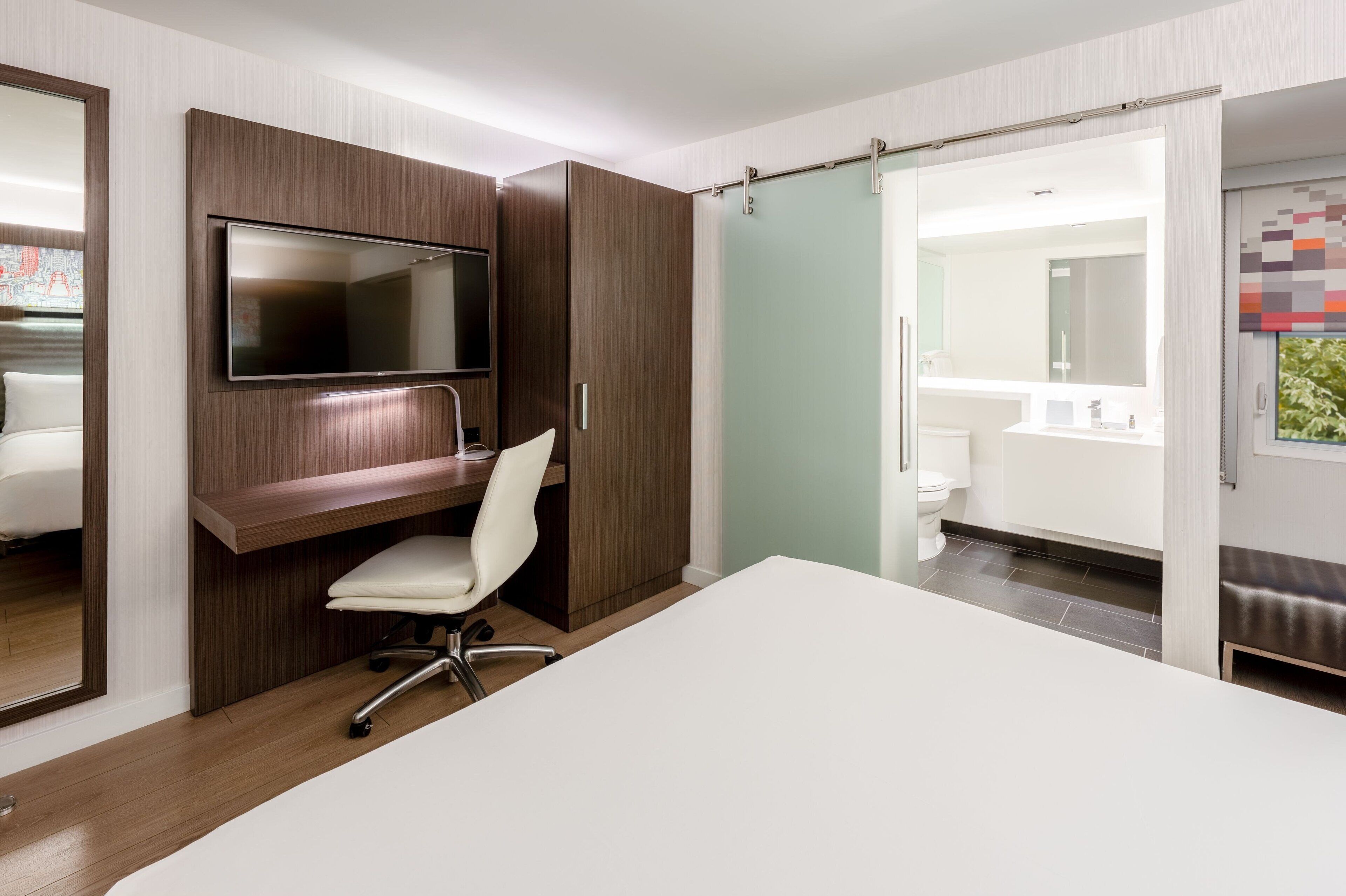 A modern hotel room with a large bed, a desk with a chair, a TV mounted on the wall, and a bathroom visible through a sliding door.