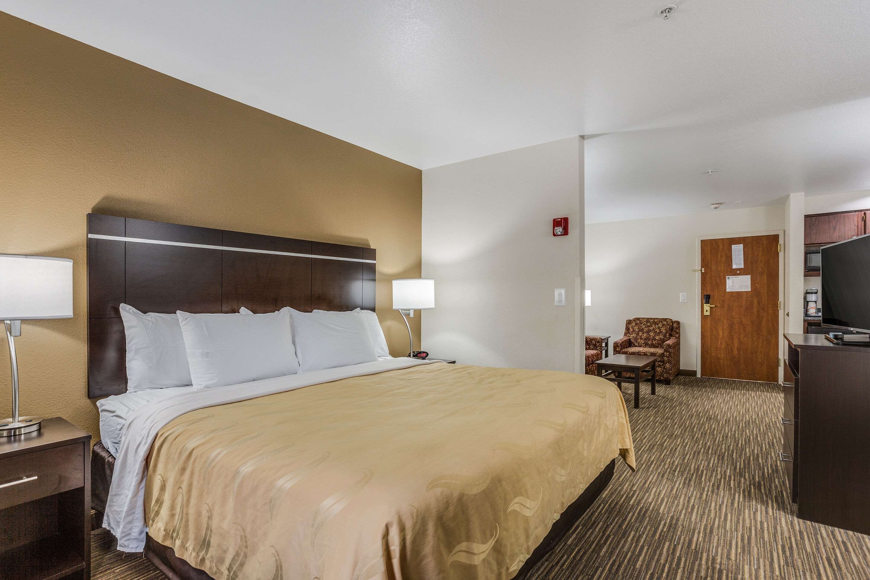 A hotel room with a large bed, bedside lamps, a desk, a chair, a television, and a door with a fire alarm.