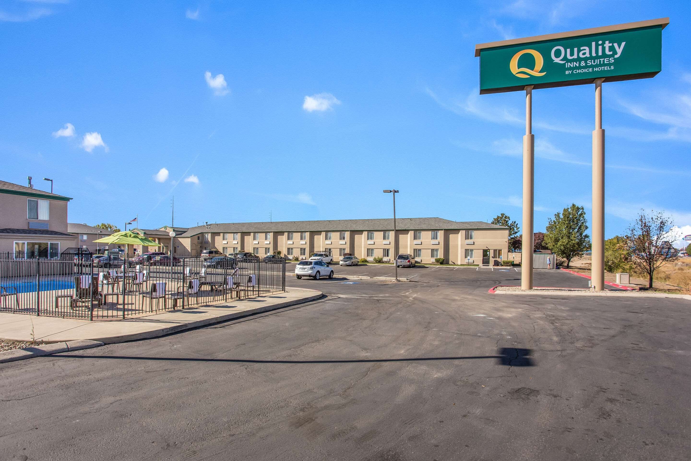 A hotel with a sign reading "Quality Inn & Suites by Choice Hotels" in front of a building with multiple parking spaces and a fenced outdoor area with chairs and umbrellas.