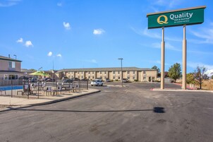 A hotel with a sign reading "Quality Inn & Suites by Choice Hotels" in front of a building with multiple parking spaces and a fenced outdoor area with chairs and umbrellas.