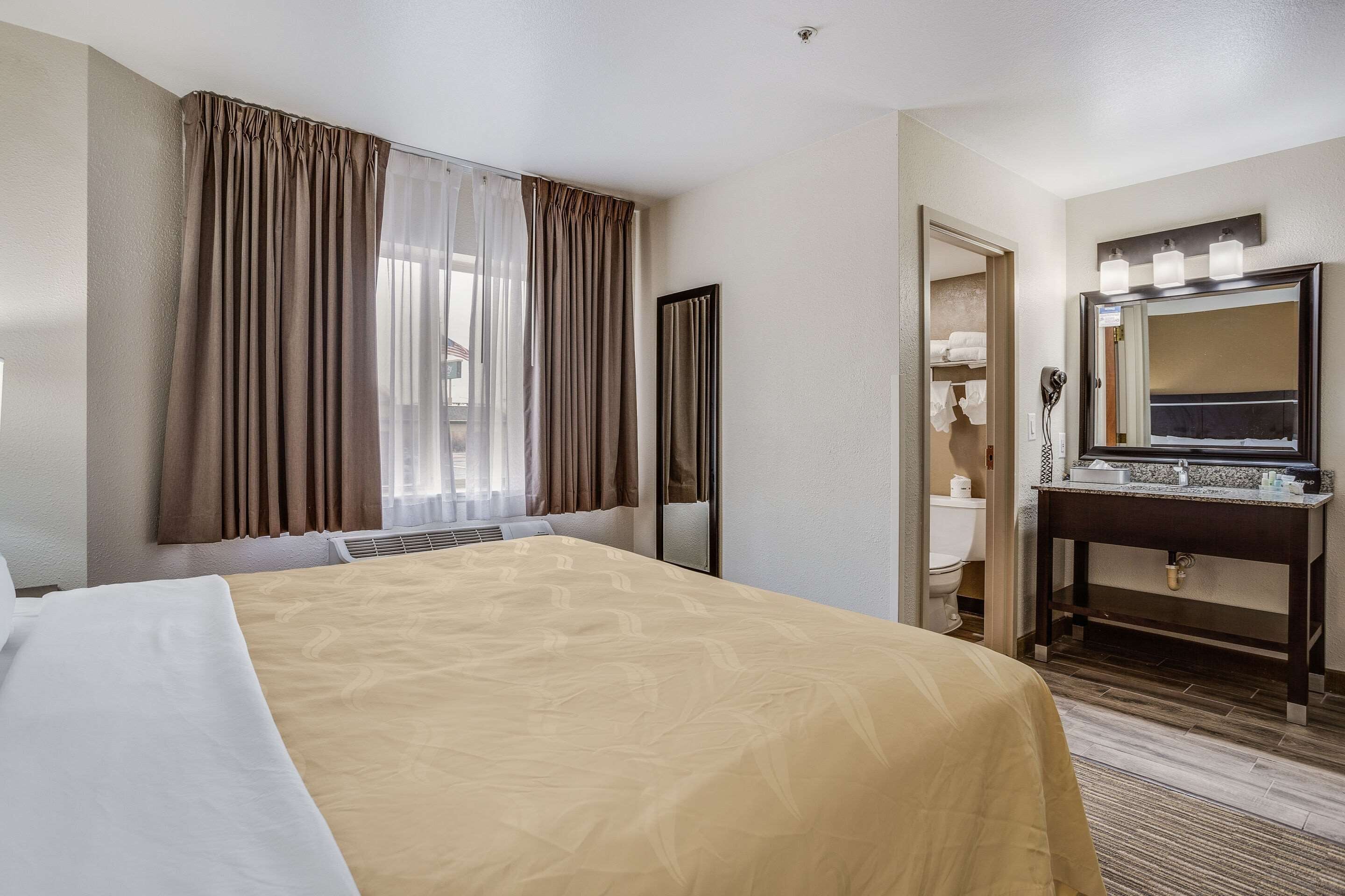 Habitación de hotel con una cama grande, una mesita de noche, un espejo y una ventana con cortinas.