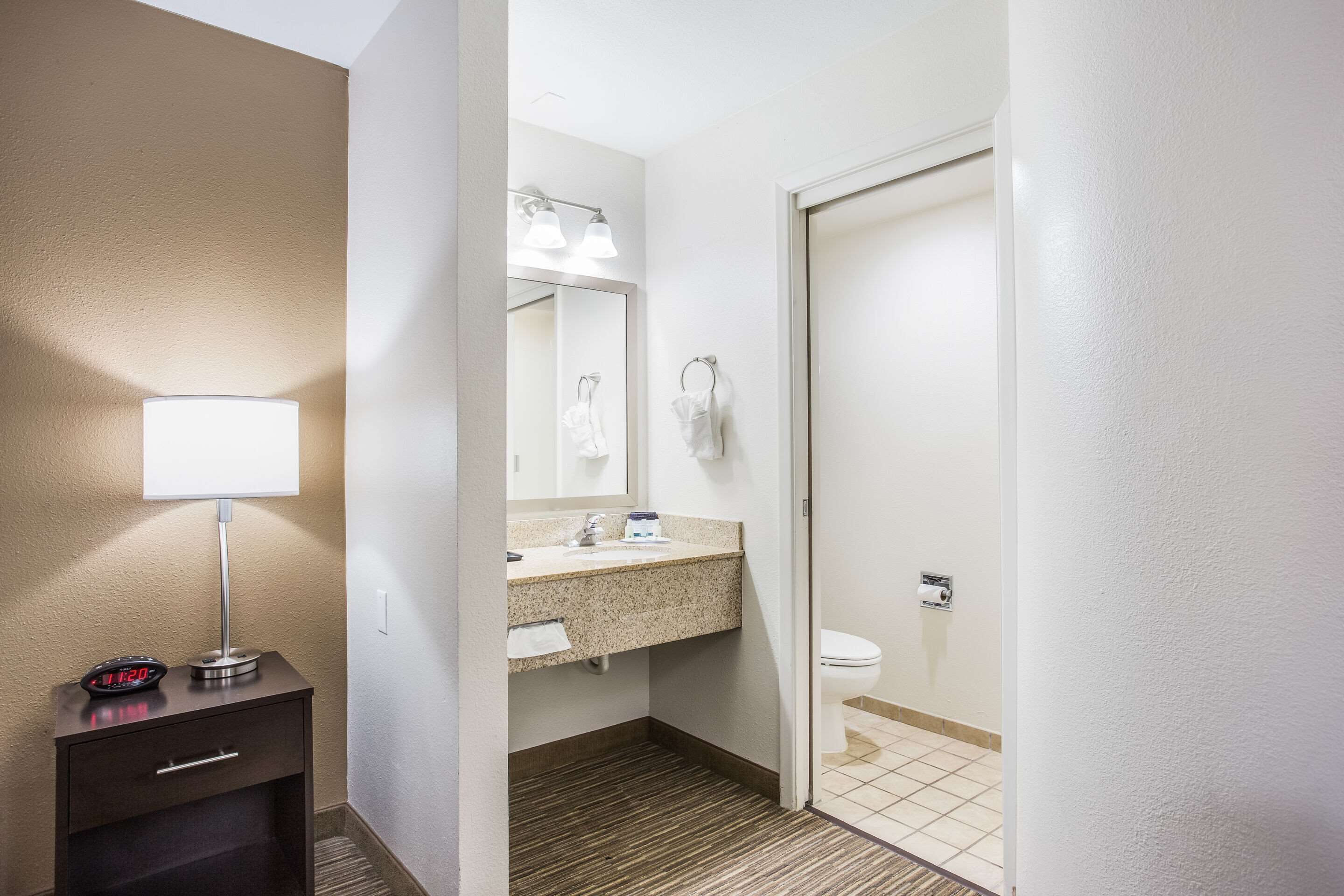 Une salle de bain d’hôtel avec un lavabo, un miroir et des toilettes. On y trouve une table de chevet avec une lampe et une horloge.