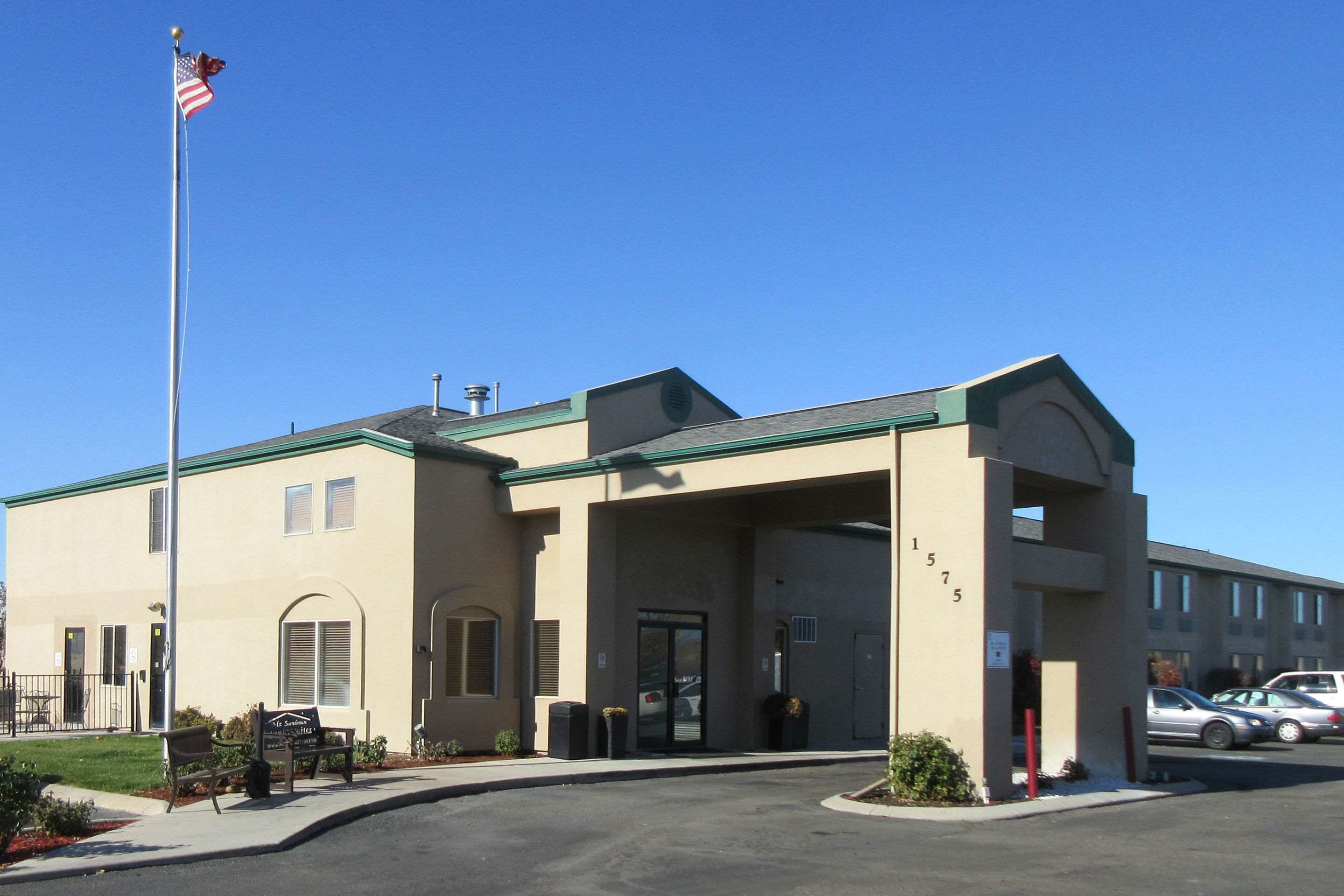 A building with a beige facade, a green roof, and a flagpole with an American flag. There are two benches outside, and a parking lot with cars.
