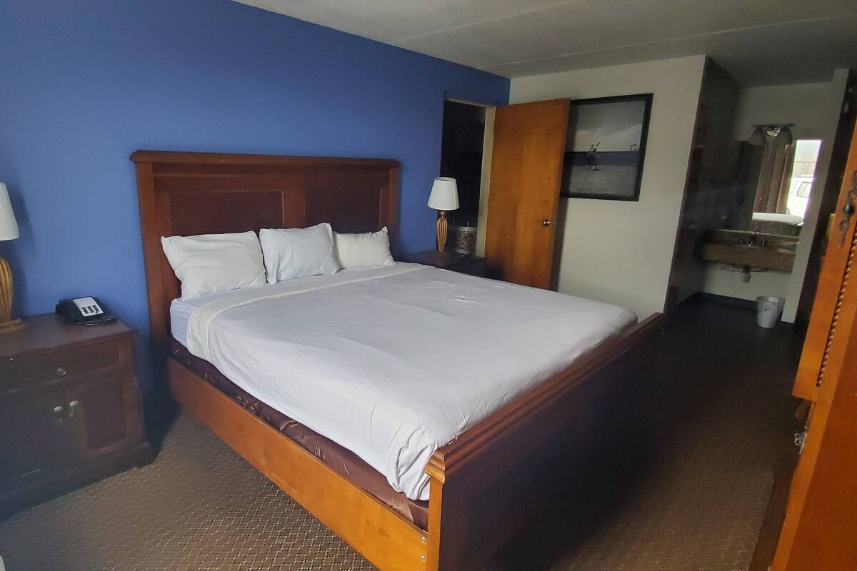 Une chambre d’hôtel avec un grand lit, une tête de lit en bois, deux tables de chevet avec des lampes, une petite table d’appoint et une salle de bain visible à travers une porte ouverte.
