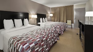 Habitación de hotel con tres camas, cada una con ropa blanca y una colcha estampada. Hay dos lámparas de noche y un escritorio con una silla.