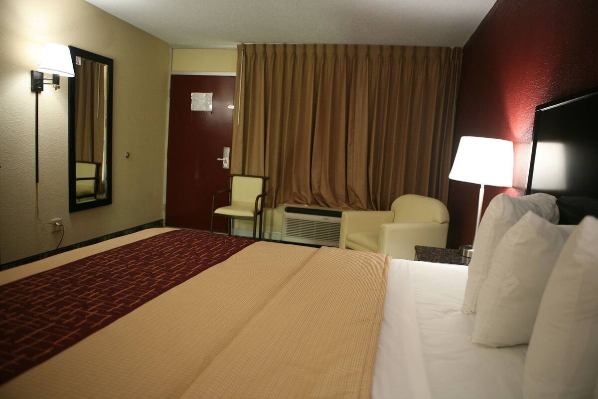 A hotel room with a bed, a chair, a desk, a lamp, a mirror, and a window with curtains.