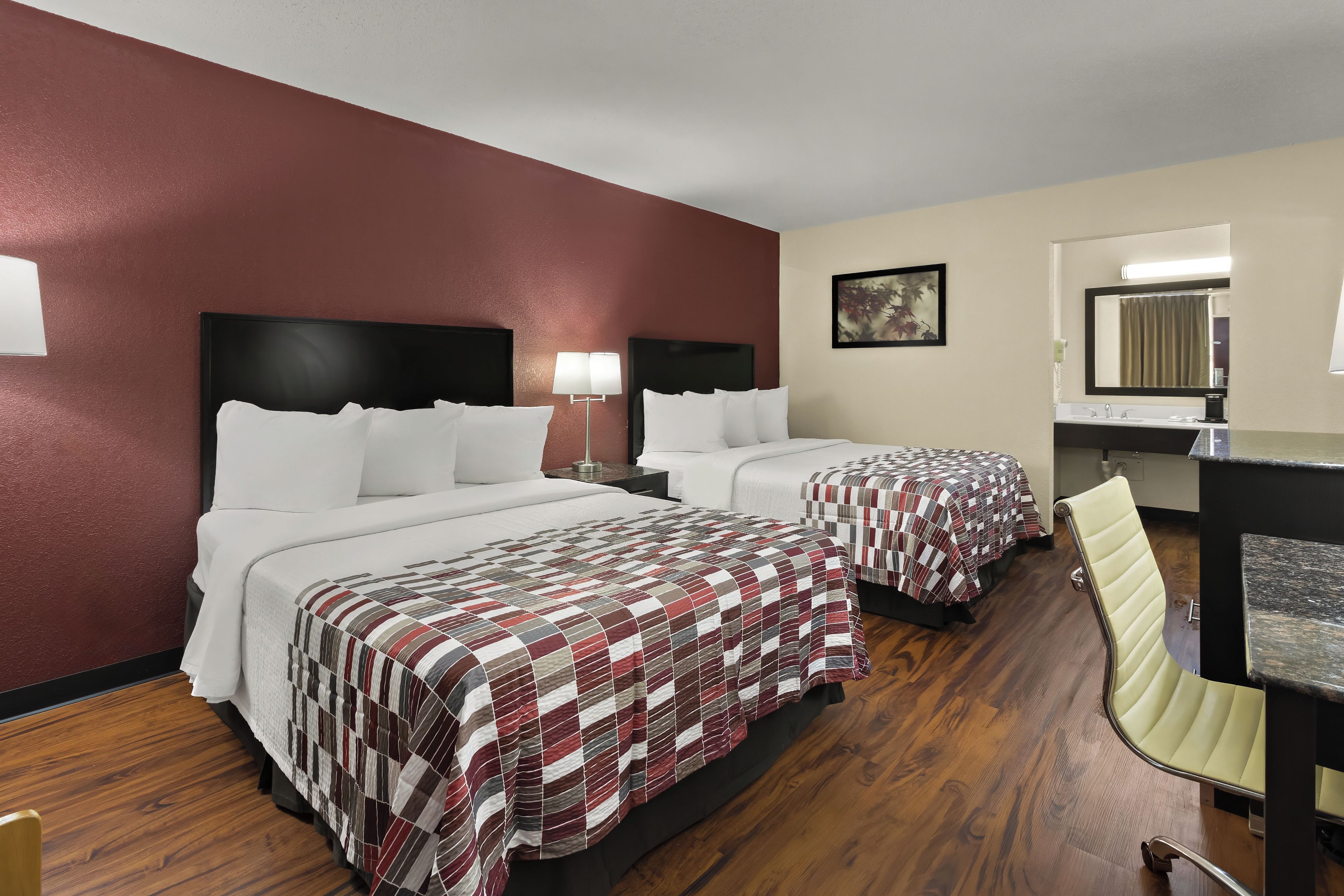 Une chambre d’hôtel avec deux lits, un bureau, une chaise, une salle de bain avec un miroir et un tableau au mur.