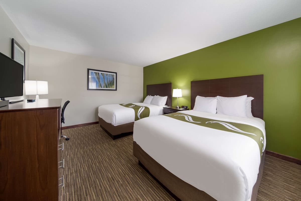 Ein Hotelzimmer mit zwei Betten, einem Schreibtisch mit Lampe, einem Fernseher und einem gerahmten Bild an der Wand.