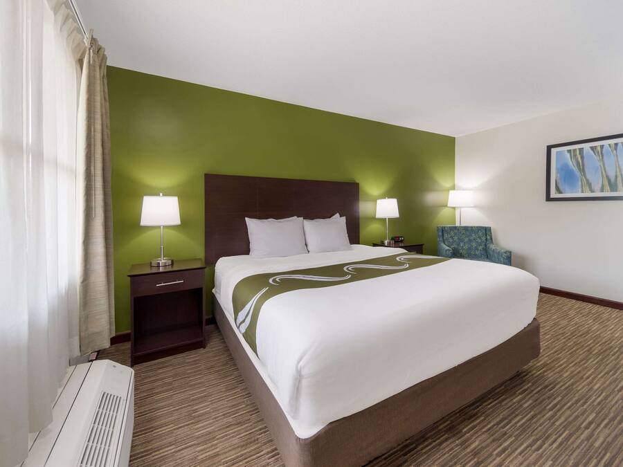 Habitació d'hotel amb un llit gran, dues taules de nit amb làmpades, una cadira, un quadre a la paret i un radiador.
