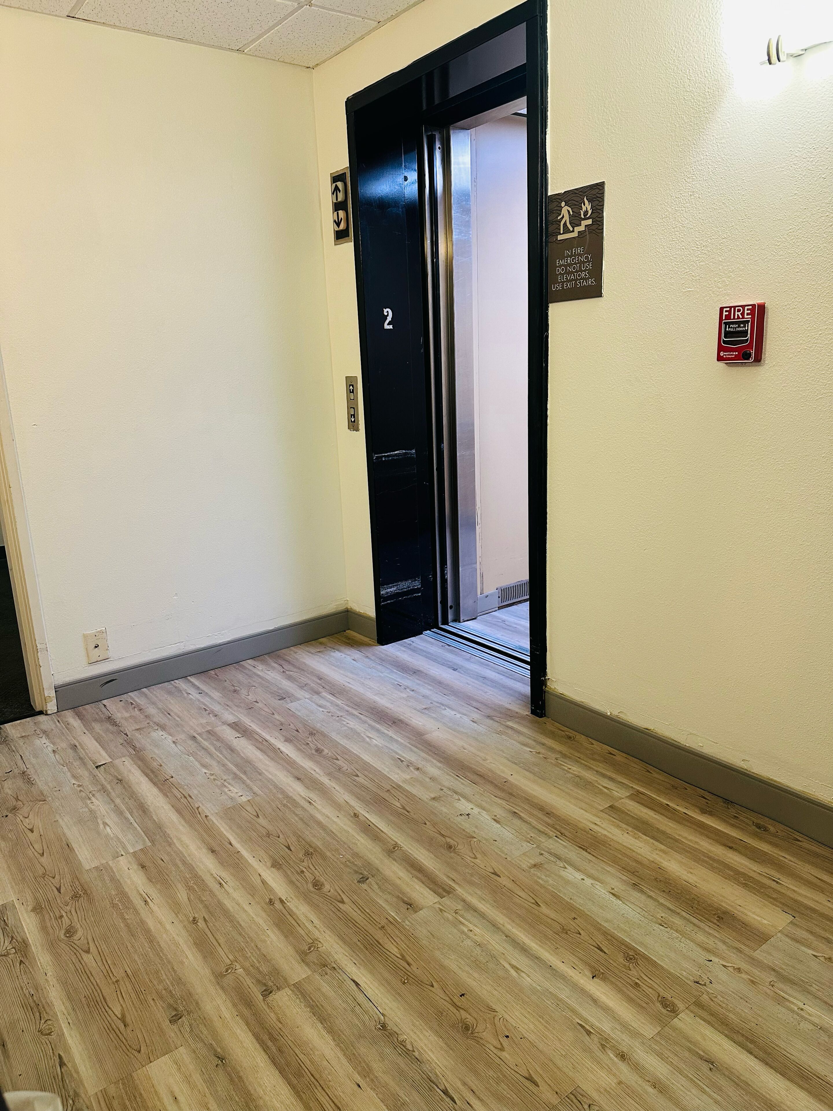 Un couloir avec un plancher en bois, une porte d’ascenseur noire portant l’inscription « 2 », un détecteur d’incendie et un panneau indiquant qu’il faut utiliser l’ascenseur en cas d’urgence.