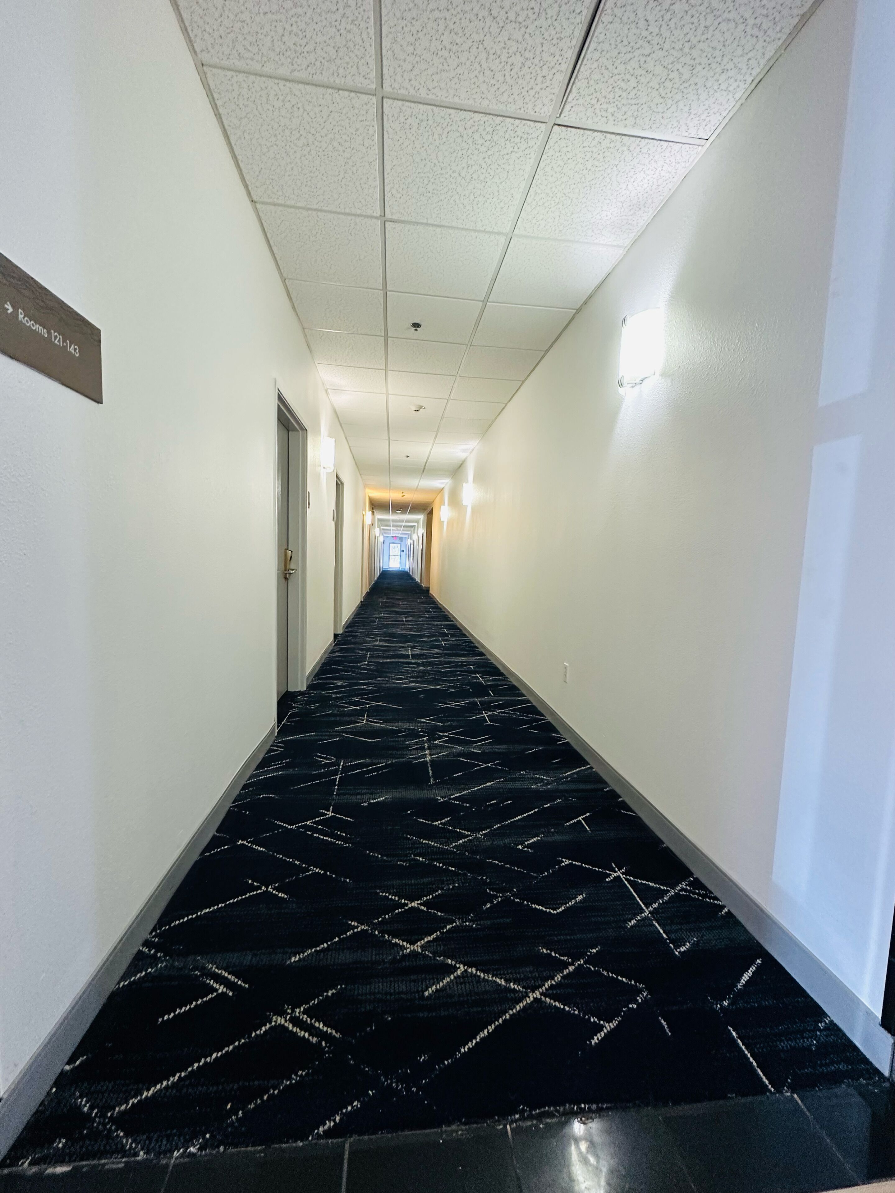 A hotel hallway with a black carpet featuring a geometric pattern, white walls, and a sign indicating rooms 121-143.