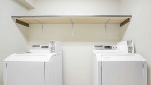 A laundry room with two white front-loading washers and dryers, a wooden shelf above, and a tiled floor. - Days Inn by Wyndham Custer (Custer)