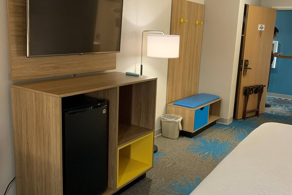 A hotel room with a bed, a TV mounted on the wall, a desk with a lamp, a small fridge, a trash bin, and a door leading to another room.