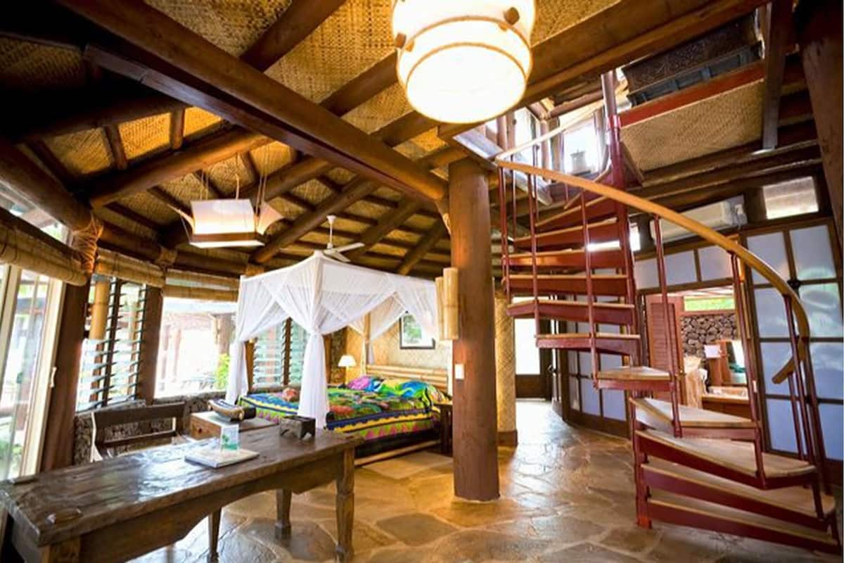 A room with a wooden ceiling, a bed with colorful bedding, a wooden table, and a spiral staircase.