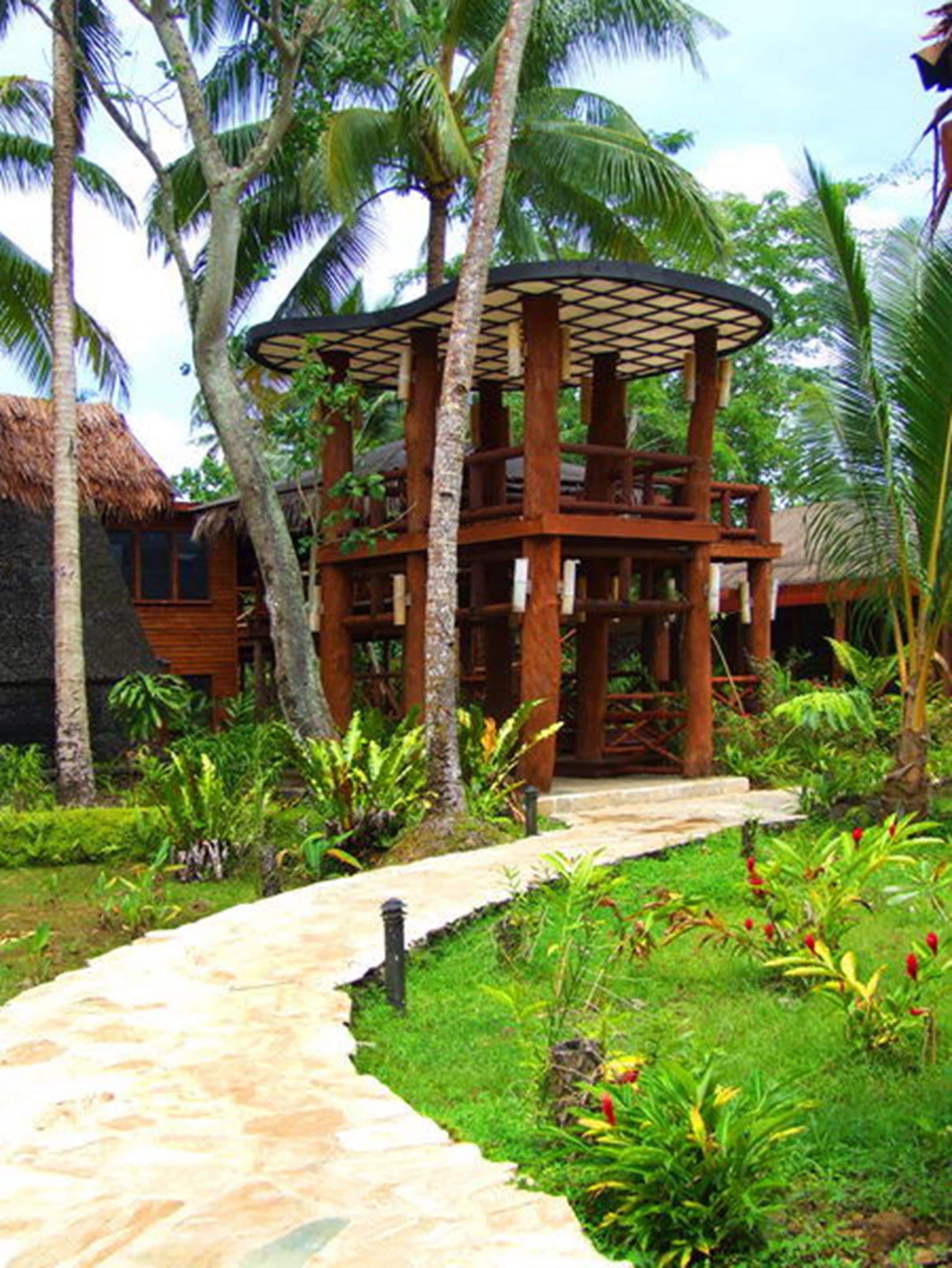 A wooden structure with a thatched roof, surrounded by lush greenery and palm trees.