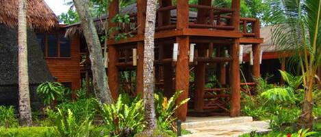 A wooden structure with a thatched roof, surrounded by lush greenery and palm trees.