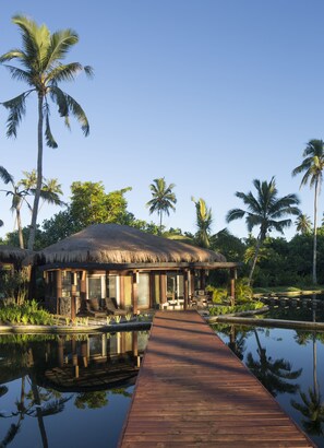 A wooden dock leading to a thatched-roof bungalow surrounded by palm trees and water.