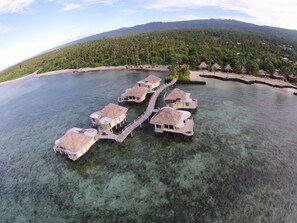 An aerial view of overwater bungalows connected by walkways, surrounded by clear water and a tropical landscape.