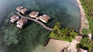 Vista aérea de bungalows sobre el agua conectados por una pasarela de madera, rodeados de aguas turquesas cristalinas y una playa de arena con palmeras.