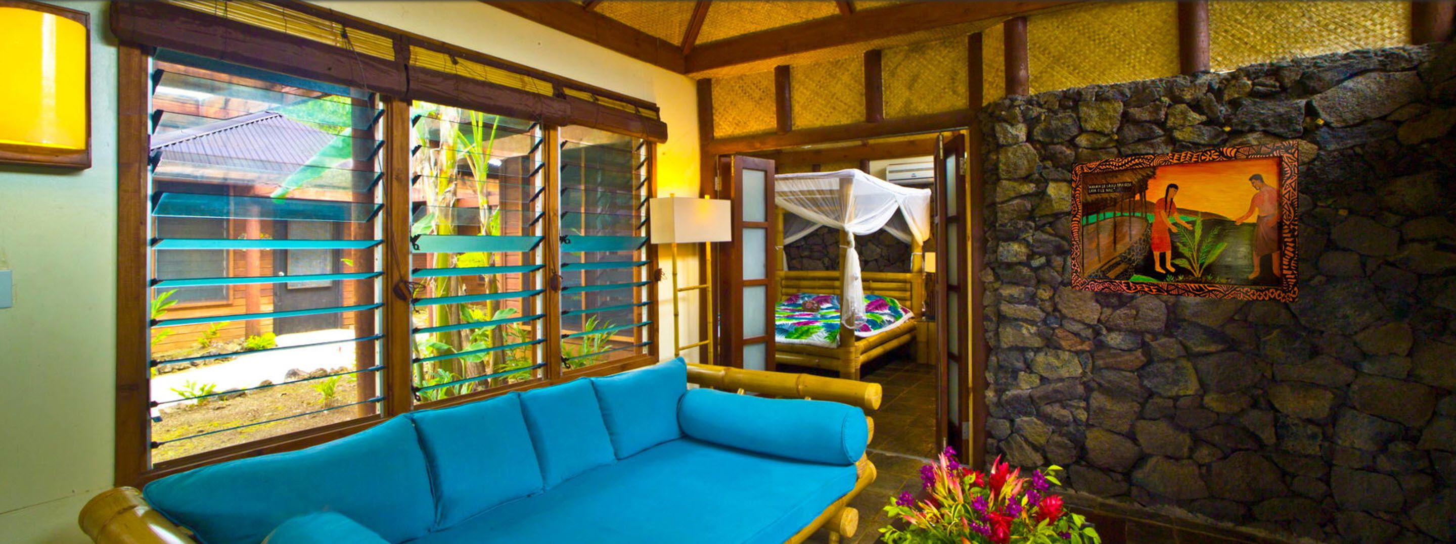 A room with a blue sofa, large windows with horizontal blinds, a stone wall, and a bed with a canopy.