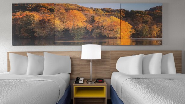 A hotel room with two beds, a nightstand with a lamp, and a large autumn-themed painting above the beds. - Days Inn by Wyndham Alma (Alma)